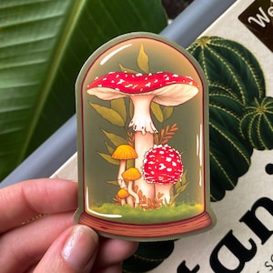 May include: A sticker of a red and white mushroom with white spots, surrounded by smaller yellow mushrooms and green foliage, inside a glass dome.