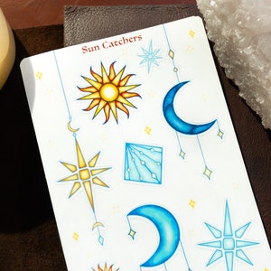 May include: A sheet of stickers with a celestial theme. The stickers feature a sun, a crescent moon, a star, and a geometric design. The stickers are all white with blue and yellow accents. The text "Sun Catchers" is printed at the top of the sheet.