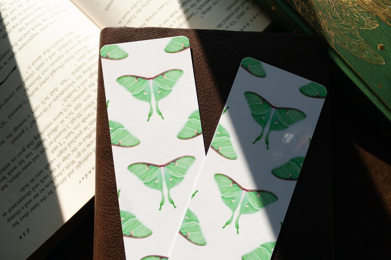 Pode incluir: Dois marcadores de p&aacute;gina brancos com um padr&atilde;o repetido de mariposas luna verdes. Os marcadores de p&aacute;gina est&atilde;o sobre uma superf&iacute;cie de couro marrom.