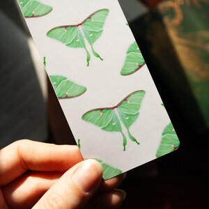 Pode incluir: Um marcador branco com um padr&atilde;o repetido de mariposas luna verdes. As mariposas t&ecirc;m um corpo verde claro e asas com uma borda verde mais escura. As asas t&ecirc;m uma mancha branca de cada lado.