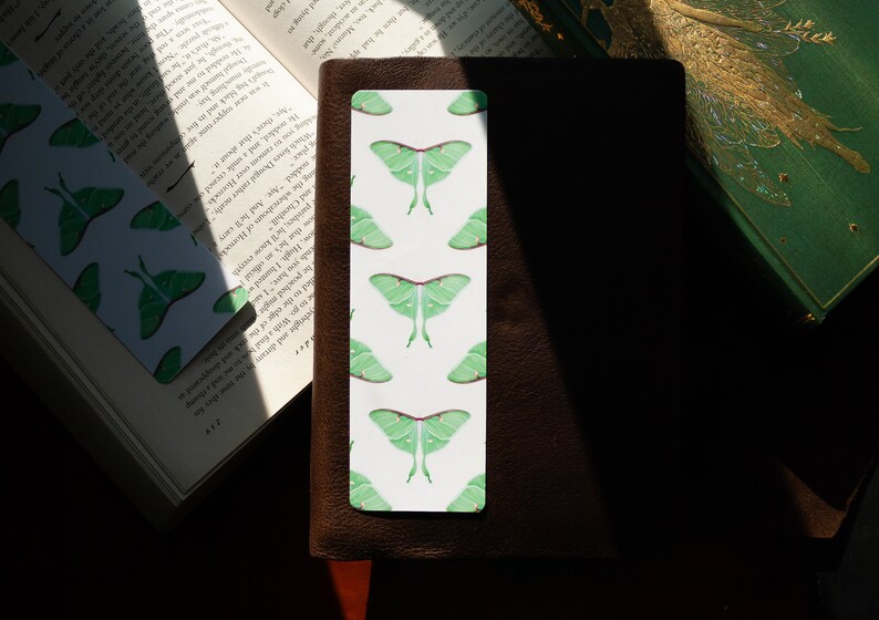 Pode incluir: Um marcador de p&aacute;gina branco com um padr&atilde;o repetido de mariposas luna verdes. O marcador est&aacute; sobre um di&aacute;rio de couro marrom.