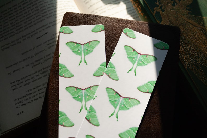 Pode incluir: Dois marcadores de p&aacute;gina brancos com um padr&atilde;o repetido de mariposas luna verdes. As mariposas s&atilde;o ilustradas em um fundo branco.