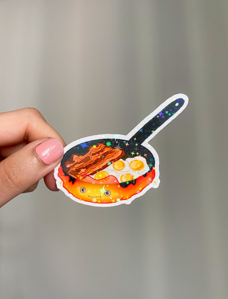 May include: A holographic sticker of a cartoon frying pan with a happy face, filled with eggs and bacon.