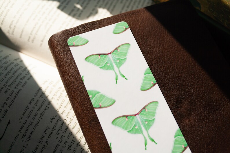 Pode incluir: Um marcador de p&aacute;gina branco com um padr&atilde;o repetido de mariposas luna verdes. As mariposas s&atilde;o ilustradas de forma realista com asas e antenas detalhadas.