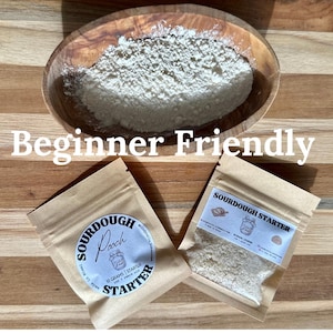 May include: A wooden bowl filled with white flour sits above two small packets of sourdough starter. The packets are labeled "Sourdough Starter" and "Beginner Friendly". The image is on a wooden surface.