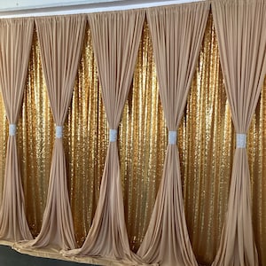 May include: A backdrop for events, featuring a gold sequin fabric with beige sheer curtains draped over it. The curtains are gathered at the top and bottom, creating a flowing effect.
