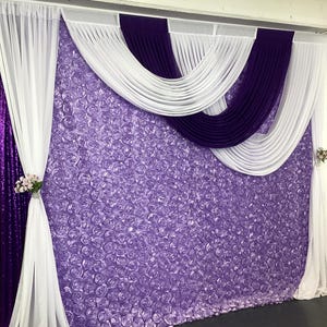May include: A backdrop for a wedding or event, featuring a purple and white fabric swag draped over a wall of lavender-colored fabric roses.