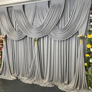 May include: A set of floor-length grey fabric curtains featuring a draped swag design across the top. The curtains are gathered at three points with gold-colored cylindrical tie-backs, each topped with small floral arrangements of roses.