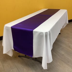 May include: A white rectangular tablecloth with a vibrant purple satin table runner. The table is set on a light brown floor, and the background is a solid yellow wall. The table is supported by a black metal frame.