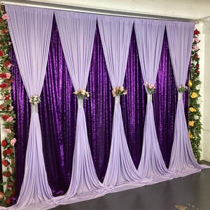 May include: A decorative backdrop featuring a purple sequined panel with draped lavender fabric. The fabric is gathered and tied with floral arrangements. The backdrop is framed with floral garlands, creating a festive and elegant setting.