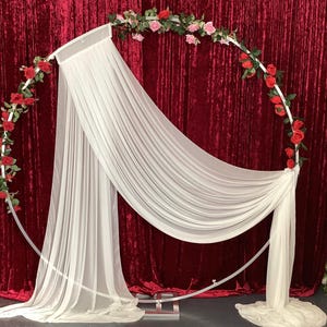 May include: A white metal wedding arch decorated with red and pink roses and draped with white fabric. The arch is set against a deep red velvet backdrop, creating a romantic and elegant setting for ceremonies.
