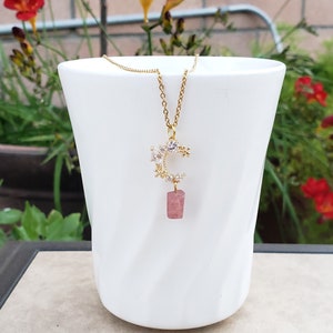 May include: A gold chain necklace with a crescent moon pendant adorned with small white crystals and a dangling pink stone.