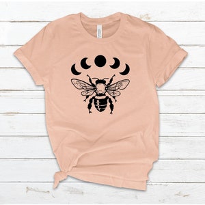 May include: A peach-colored t-shirt with a black bee and crescent moon design. The bee is centered on the shirt and the moons are arranged in a semi-circle above the bee.