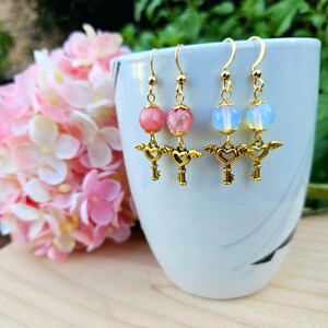 May include: A pair of gold earrings with a heart-shaped key charm and a pink or blue gemstone bead. The earrings are hanging from a white mug.