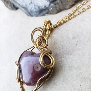 May include: A gold wire wrapped pendant featuring a heart-shaped purple agate stone with white markings. The pendant is attached to a gold chain.
