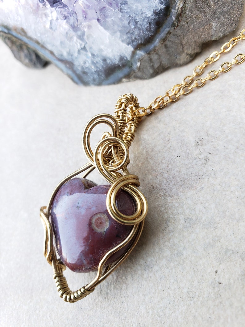 May include: A gold wire wrapped pendant featuring a heart-shaped purple agate stone with white markings. The pendant is attached to a gold chain.