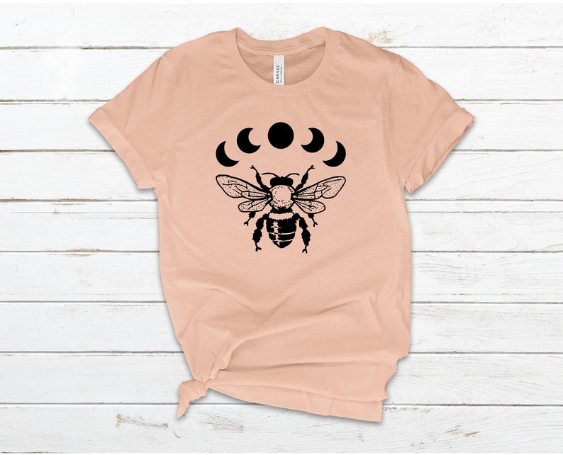 May include: A peach-colored t-shirt with a black bee and crescent moon design. The bee is centered on the shirt and the moons are arranged in a semi-circle above the bee.