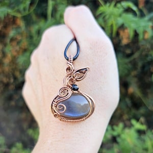 May include: A handcrafted copper wire pendant with a blue-gray stone and black bead accent. The pendant has an intricate, swirling design and hangs from a black cord. The background is blurred green foliage.