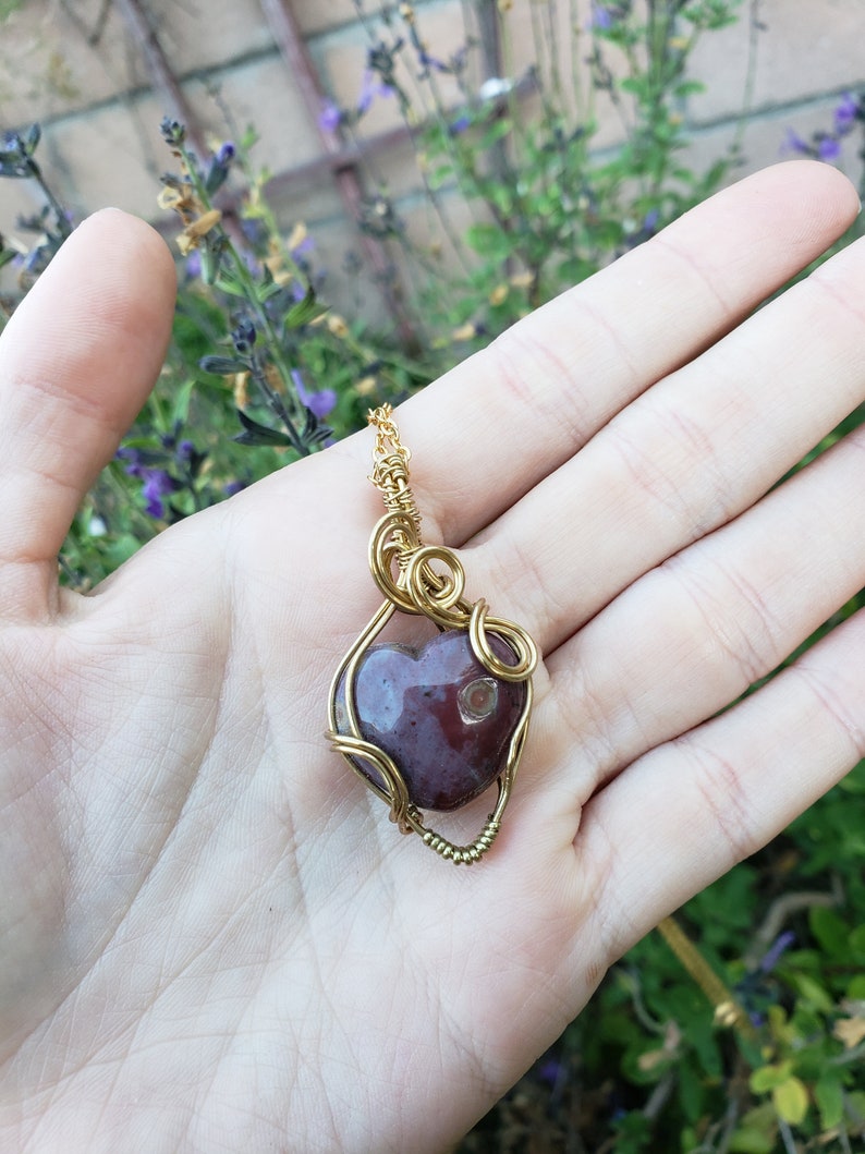 May include: A heart-shaped pendant made of red agate wrapped in gold wire. The pendant is hanging from a gold chain.