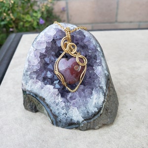 May include: A gold wire wrapped heart-shaped pendant with a red and brown agate stone, displayed on a purple amethyst geode.