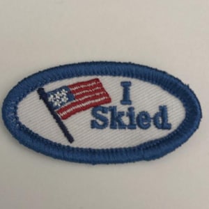 May include: Oval-shaped embroidered patch with a blue border and the words "I Skied" in blue. The patch features a small American flag with red and white stripes and blue stars.