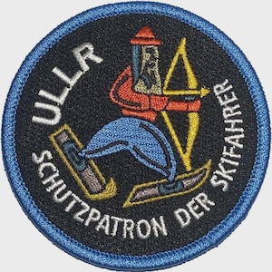 Peut inclure: Écusson brodé circulaire avec un fond noir et une bordure bleue. Il représente un skieur en rouge et bleu, tenant un arc et une flèche jaunes. Le texte en blanc autour du bord indique "ULLR SCHUTZPATRON DER SKIFAHRER".