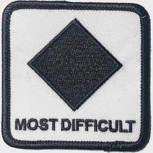 May include: Embroidered square patch featuring a black diamond on a white background. The words "MOST DIFFICULT" are stitched in black beneath the diamond. The patch is edged with a black border.