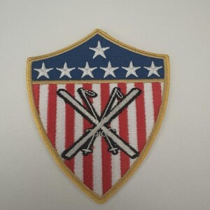 Pode incluir: Remendo bordado em forma de escudo. A seção superior é azul com seis estrelas brancas. O corpo principal tem listras verticais vermelhas e brancas. Dois esquis cruzados estão no centro, com uma borda dourada.