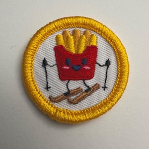 Puede incluir: Parche bordado con un contenedor de patatas fritas rojo sonriente con patatas fritas, esquiando sobre esquís marrones. El parche tiene bastones de esquí negros y un borde amarillo, sobre un fondo blanco.