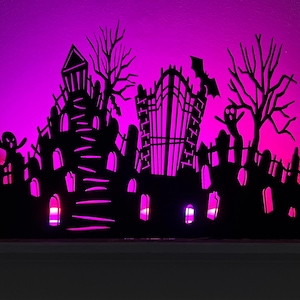 May include: A black Halloween-themed silhouette featuring a haunted house, graveyard, and bare trees, illuminated by a vibrant pink glow. The decorative piece includes a bat and ghost, creating a spooky ambiance for seasonal decor.