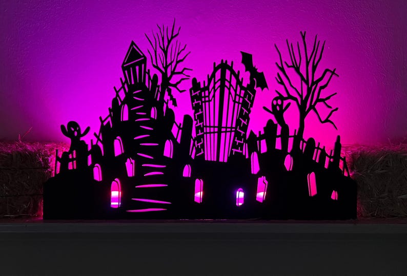May include: A black Halloween-themed silhouette featuring a haunted house, graveyard, and bare trees, illuminated by a vibrant pink glow. The decorative piece includes a bat and ghost, creating a spooky ambiance for seasonal decor.