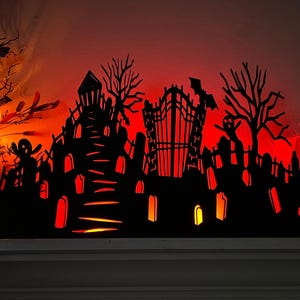 May include: A black Halloween-themed silhouette featuring a haunted house, graveyard, and trees, illuminated with a warm orange glow. A bat is flying above a gate. The decorative piece is perfect for Halloween.