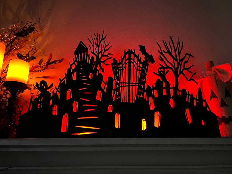 May include: A black Halloween-themed silhouette featuring a haunted house, graveyard, and trees, illuminated with a warm orange glow. A bat is flying above a gate. The decorative piece is perfect for Halloween.