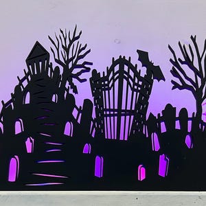 May include: A black Halloween-themed silhouette featuring a graveyard, haunted house, and bat. The scene is illuminated with purple lights, creating a spooky ambiance. Hay bales flank the sides, enhancing the festive decor.