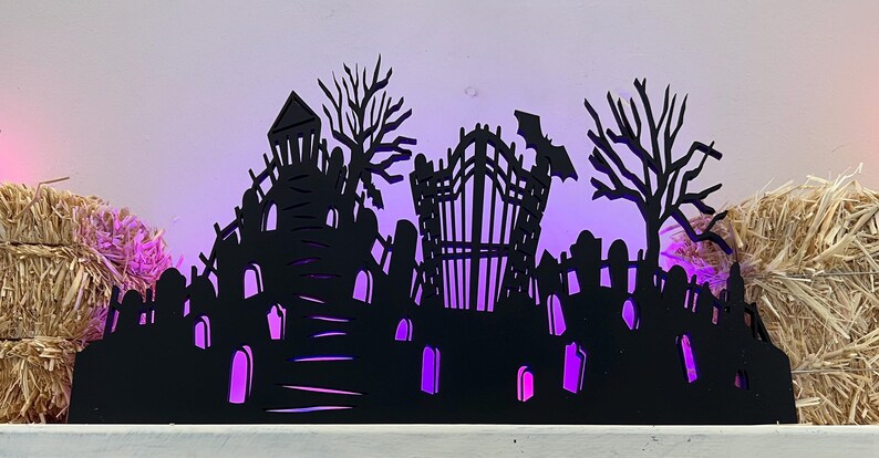 May include: A black Halloween-themed silhouette featuring a graveyard, haunted house, and bat. The scene is illuminated with purple lights, creating a spooky ambiance. Hay bales flank the sides, enhancing the festive decor.