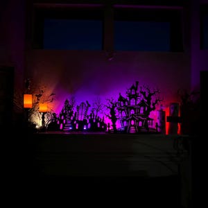 May include: A Halloween-themed display featuring a silhouette of a haunted house, graveyard, and trees, illuminated with purple, orange, and red lights. The scene is set on a shelf, creating a spooky ambiance.