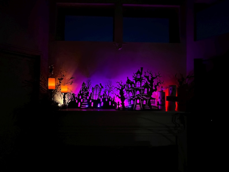 May include: A Halloween-themed display featuring a silhouette of a haunted house, graveyard, and trees, illuminated with purple, orange, and red lights. The scene is set on a shelf, creating a spooky ambiance.