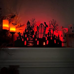 May include: A Halloween-themed display featuring a black silhouette of a haunted town with a graveyard, lit with red light. Two orange candles and a pumpkin with the word "BOO" are also visible.