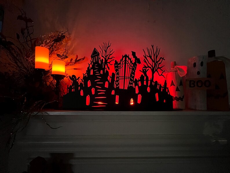 May include: A Halloween-themed display featuring a black silhouette of a haunted town with a graveyard, lit with red light. Two orange candles and a pumpkin with the word "BOO" are also visible.