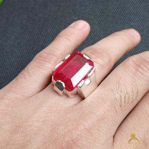 Puede incluir: Un anillo de plata con una gran gema rectangular de color rojo rub&iacute;. El anillo tiene una banda ancha y detalles decorativos en los lados del engaste de la gema. El anillo se muestra en una mano.