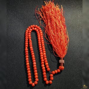 May include: A strand of red beads with a large tassel. The beads are a vibrant coral color and are strung together. The tassel is made of red threads and is attached to the end of the strand. The background is a dark, textured surface.