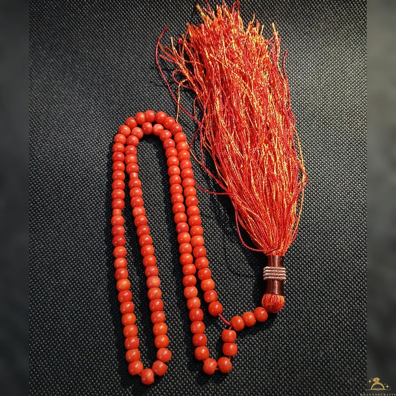 May include: A strand of red beads with a large tassel. The beads are a vibrant coral color and are strung together. The tassel is made of red threads and is attached to the end of the strand. The background is a dark, textured surface.