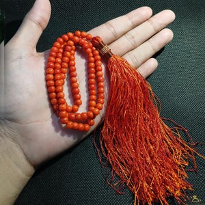 May include: A vibrant orange beaded necklace with a long, matching tassel. The necklace is composed of small, round beads. The tassel is made of numerous strands of orange thread, creating a flowing effect. The necklace is displayed on a hand.