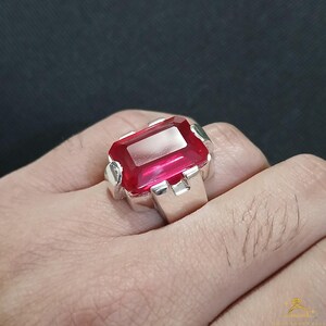 Puede incluir: Un anillo de plata con una gran gema roja rectangular y vibrante. La gema est&aacute; engastada en una banda de plata con un dise&ntilde;o sencillo y moderno. El anillo se muestra en una mano sobre un fondo oscuro.