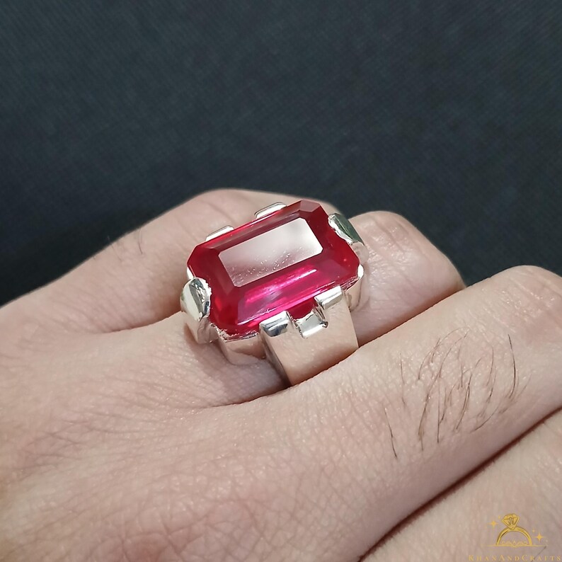 Puede incluir: Un anillo de plata con una gran gema roja rectangular y vibrante. La gema est&aacute; engastada en una banda de plata con un dise&ntilde;o sencillo y moderno. El anillo se muestra en una mano sobre un fondo oscuro.