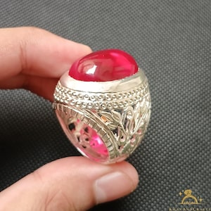 May include: A silver ring with a large, oval, bright red gemstone. The ring has intricate floral and scrollwork designs on the band and around the gemstone setting. The gemstone is smooth and polished.