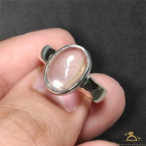 May include: A silver ring featuring a smooth, oval-shaped, translucent light pink gemstone set in a polished metal bezel. The stone displays subtle internal inclusions and a reflective surface, mounted on a simple, solid silver band.
