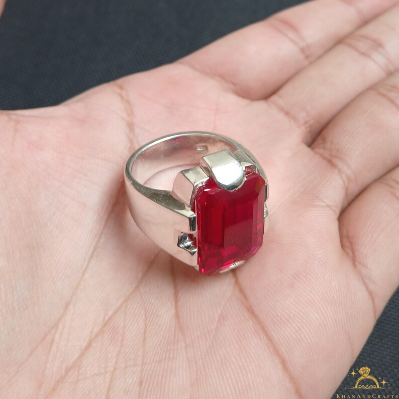 Puede incluir: Un anillo de plata con una gran gema roja brillante de forma rectangular. El anillo tiene una banda gruesa y un dise&ntilde;o geom&eacute;trico moderno alrededor de la piedra. El anillo se muestra en una mano.