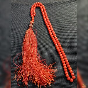 May include: A red prayer bead necklace with a long tassel. The beads are small and round, and the tassel is made of many strands of red and orange thread. The necklace is displayed on a dark gray surface.