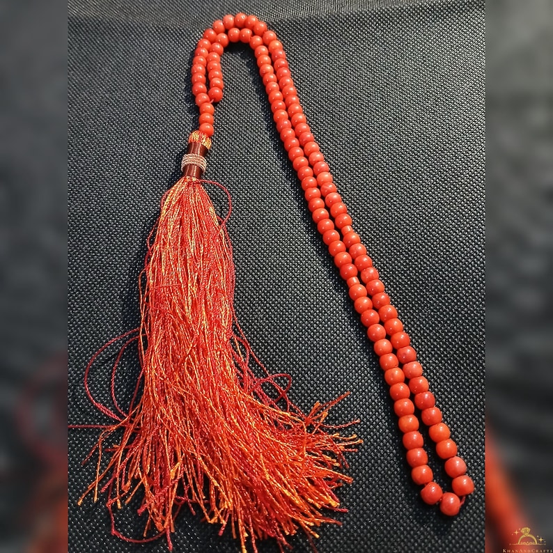 May include: A red prayer bead necklace with a long tassel. The beads are small and round, and the tassel is made of many strands of red and orange thread. The necklace is displayed on a dark gray surface.